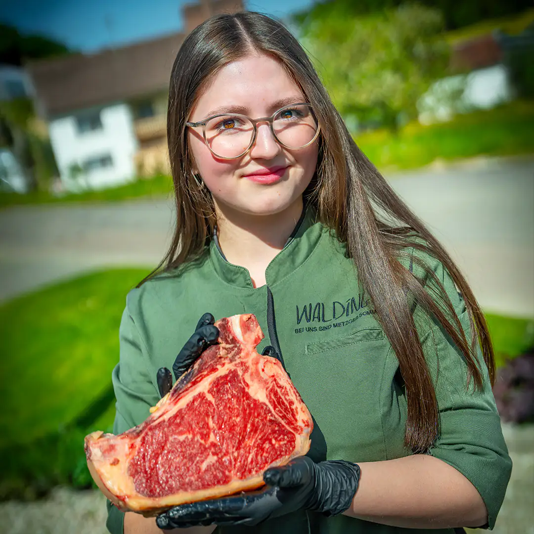 Junge Metzgerin in grüner Waldinger-Arbeitskleidung hält ein saftiges T-Bone-Steak mit schwarzen Handschuhen.